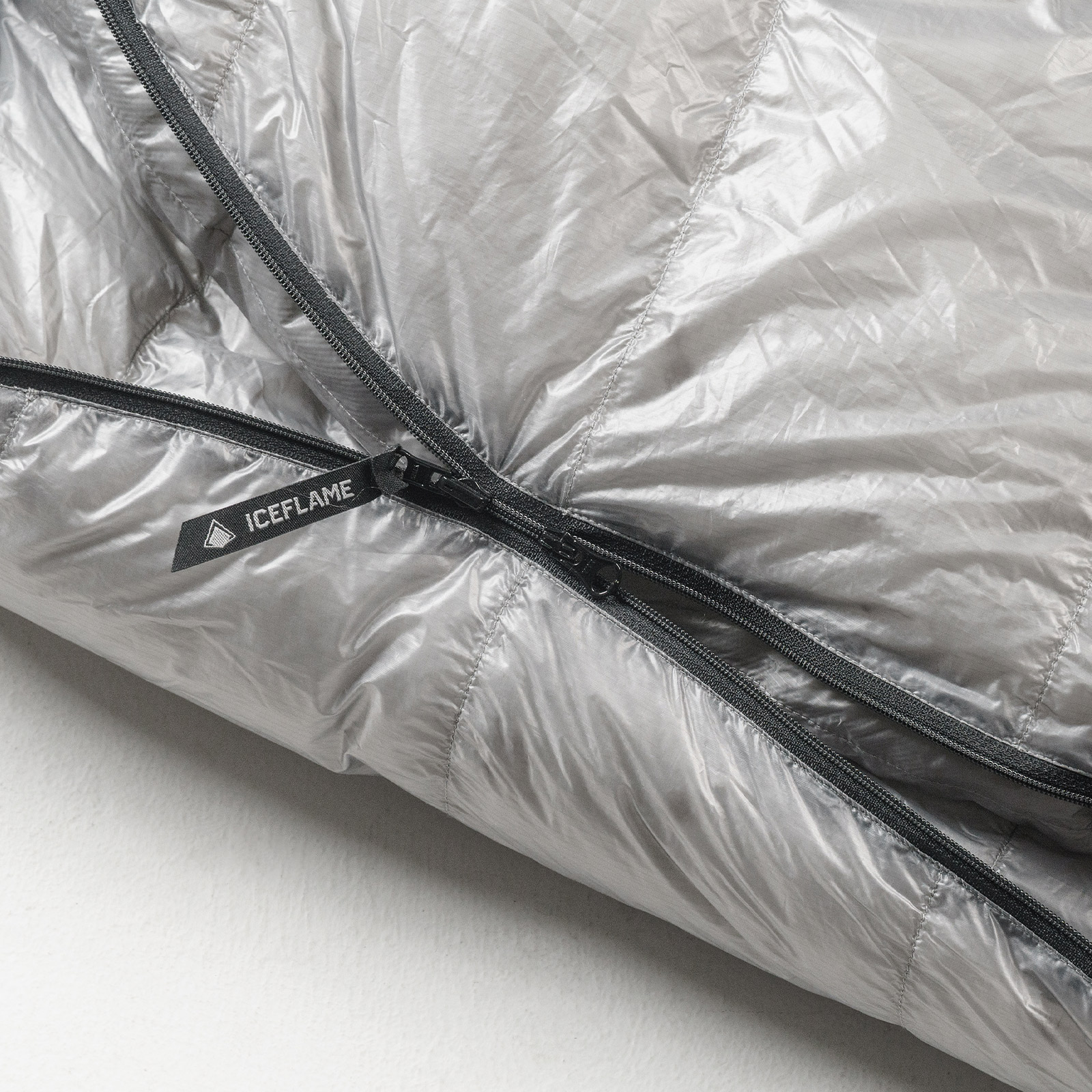 Close-up of a silver puffer jacket with a black IceFlame zipper pull and seams visible along the fabric edge.