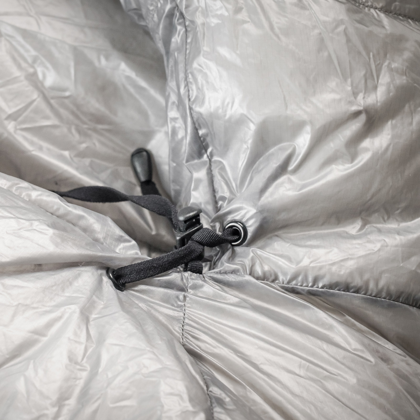 Close-up of a shiny silver quilted fabric with black drawcords and a plastic cord lock/stopper attached to a metal grommeted eyelet.