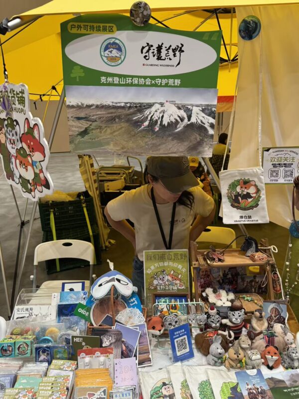 Market stall with yellow canopy promoting wilderness conservation; banner shows Chinese text and a snowy mountain map, person in hat behind table of plush animals and souvenirs.