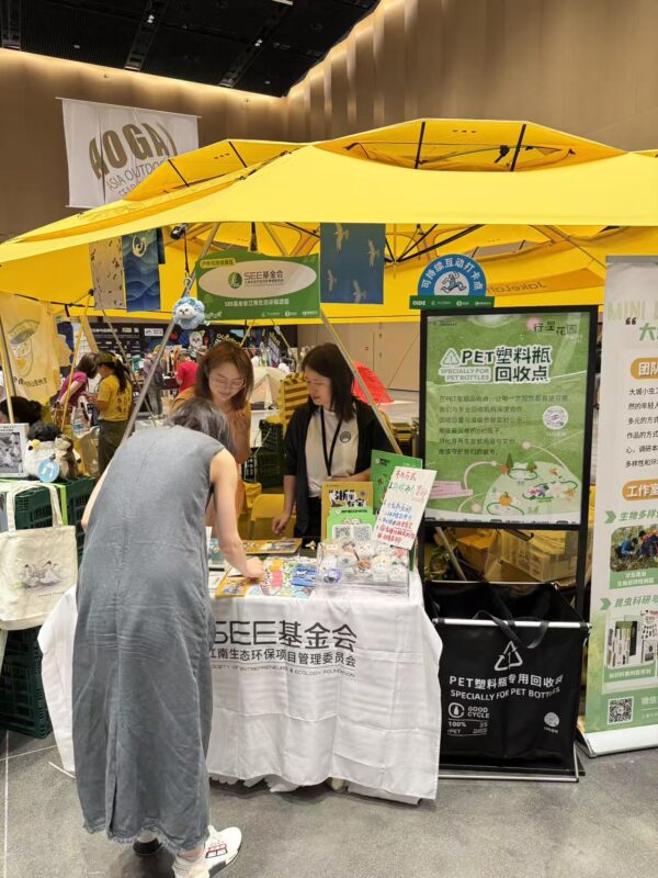 Exhibition booth under yellow canopy with posters about pet bottle recycling and ecological projects; people browse items on a table in a convention hall.