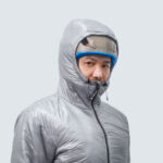 Person wearing a silver insulated puffer jacket with hood up, looking at the camera against a pale background.